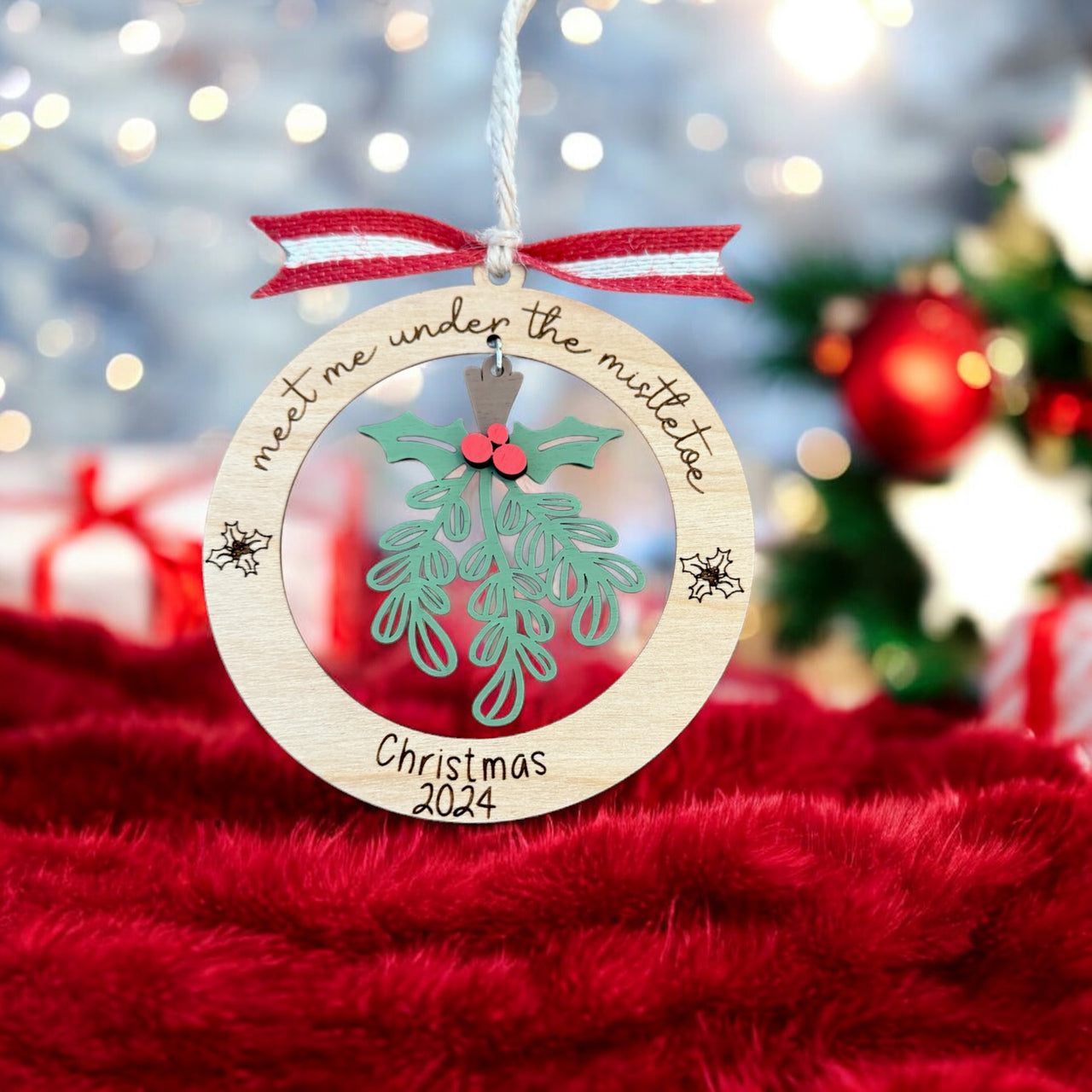 A wooden ornament with a Christmas tree and mistletoe hangs on a string, with a red ribbon and the text "Meet me under the mistletoe" written on it.