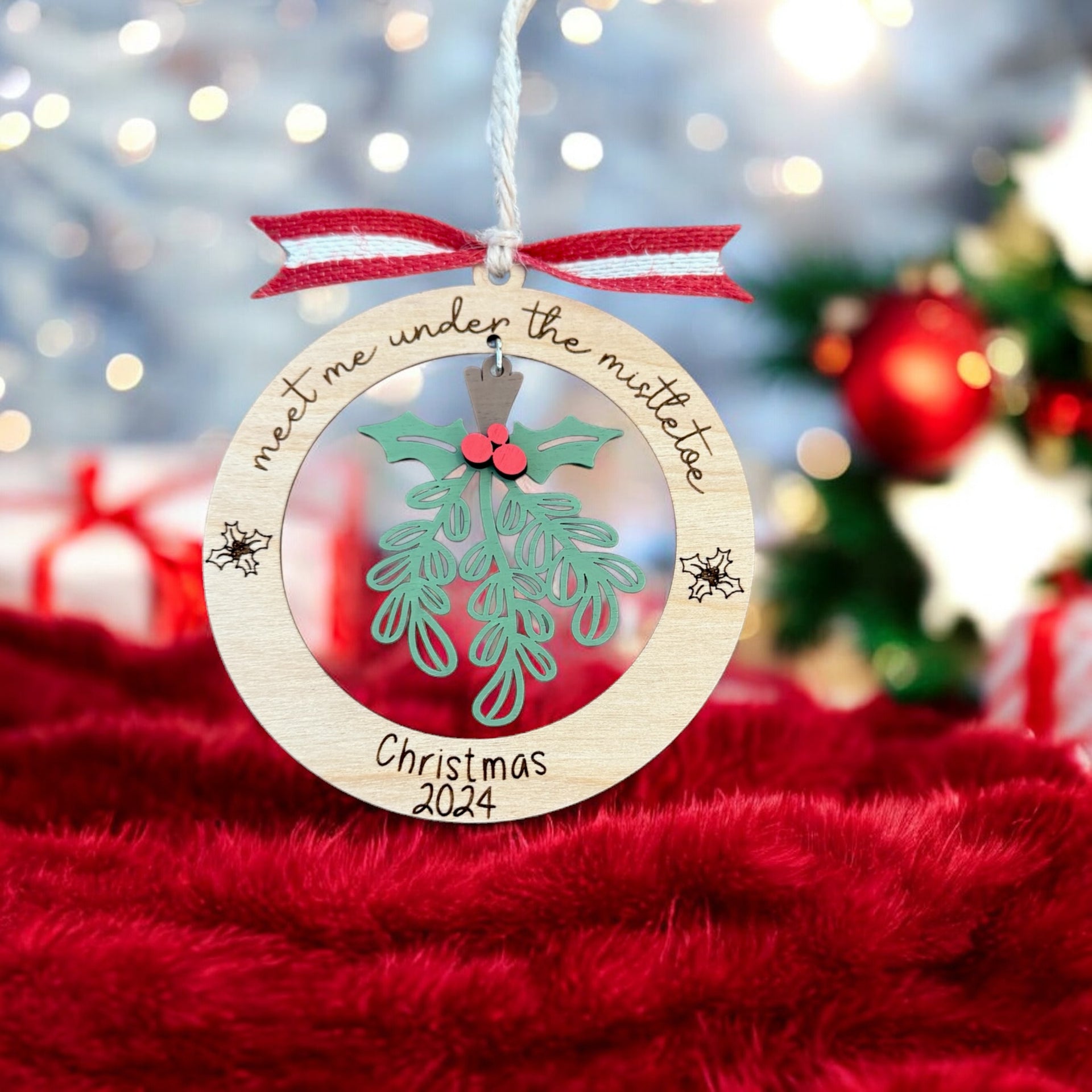 A wooden ornament with a Christmas tree and mistletoe hangs on a string, with a red ribbon and the text "Meet me under the mistletoe" written on it.