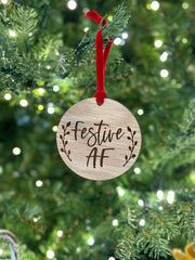 A wooden ornament with the text "Festive AF" engraved on it, hanging from a tree branch with a red ribbon.