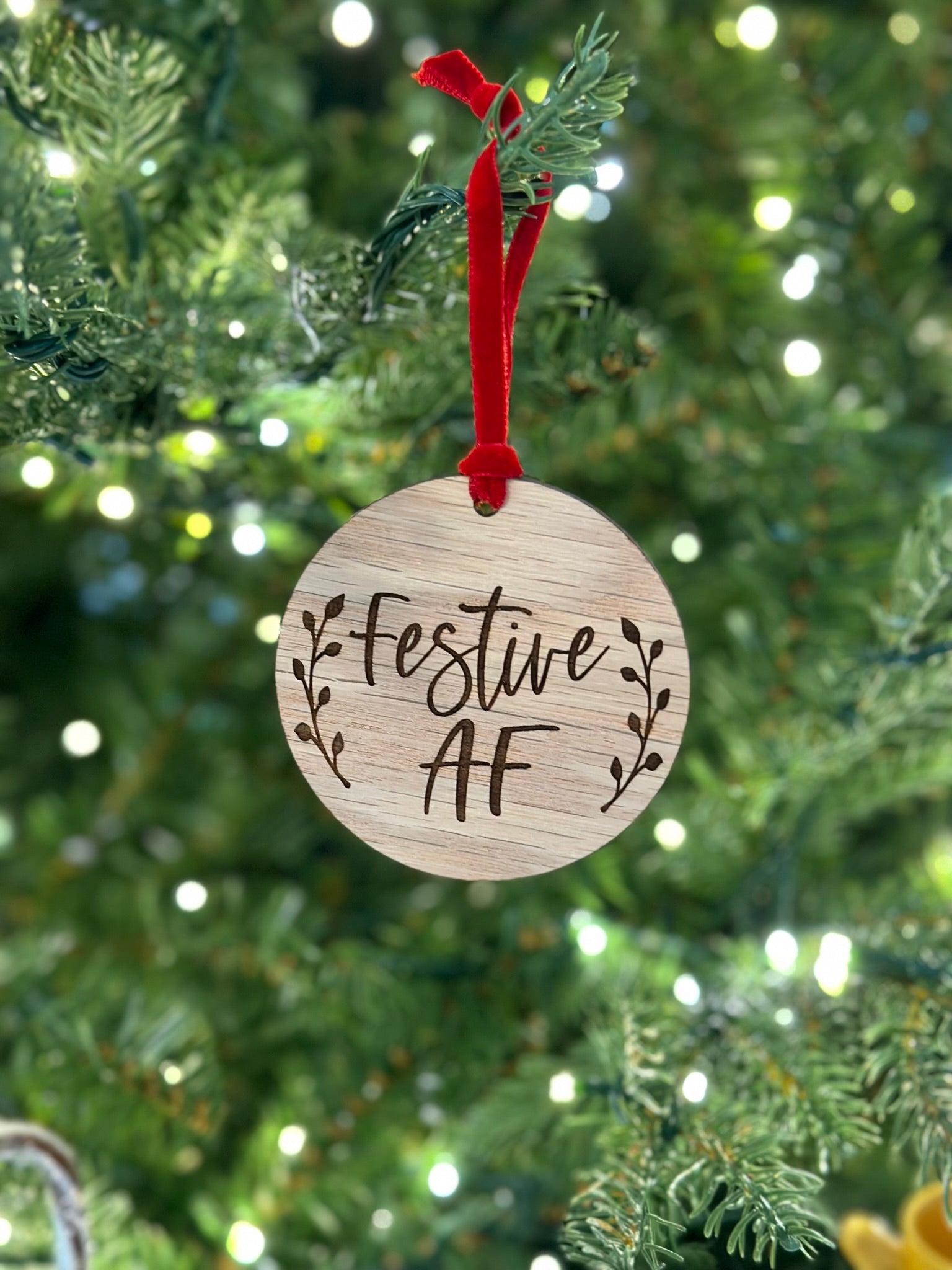 A wooden ornament with the text "Festive AF" engraved on it, hanging from a tree branch with a red ribbon.