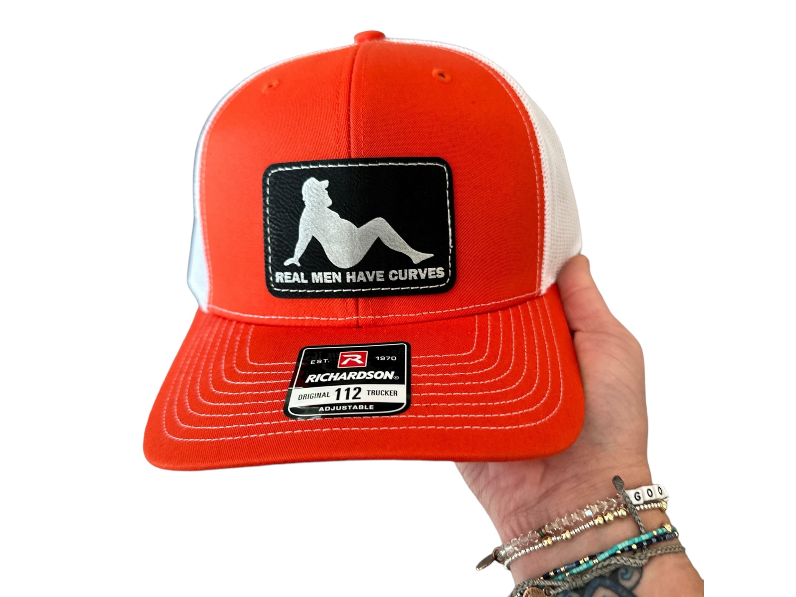 A person is holding a red baseball cap with a patch on it that says "Real men have curves".