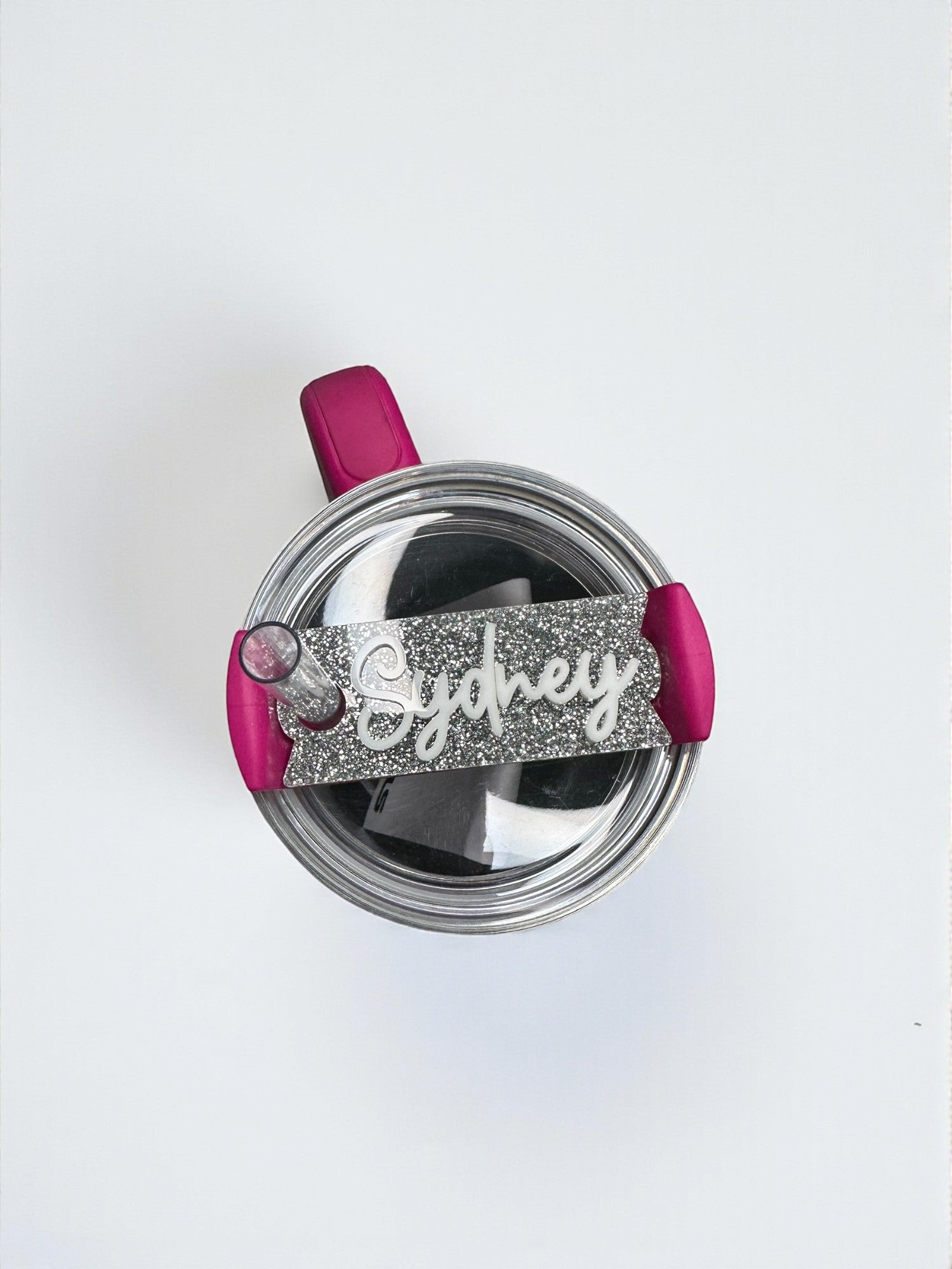 A pink and silver personalized name tag with the name "Sydney" is attached to a clear glass container with a pink handle.